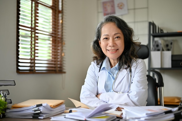 Profesjonalna starsza azjatycka lekarka przy biurku uśmiecha się i patrzy w kamerę
