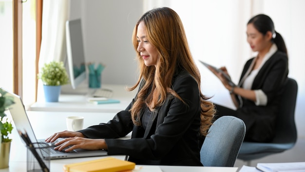 Profesjonalne azjatyckie businesswoman siedzi przy biurku i pracuje na swoim laptopie.