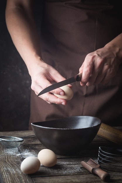 Profesjonalne ręce szefa kuchni rozbijają jajko do miski, aby zrobić ciasto na drewnianym stole, w ciemności