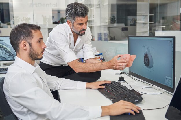 Profesjonalni inżynierowie płci męskiej obserwujący respirator podczas projektowania modelu 3D maski na komputerze w biurze modelowym podczas epidemii koronaawirusa