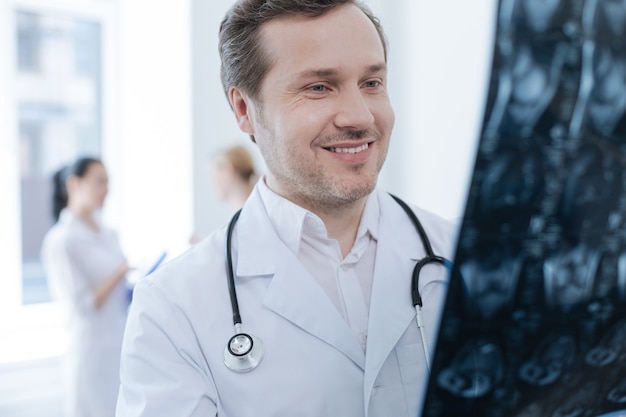 Profesjonalny brodaty uśmiechnięty neurochirurg pracuje w klinice i trzyma zdjęcie rentgenowskie, podczas gdy jego koledzy cieszą się rozmową za plecami