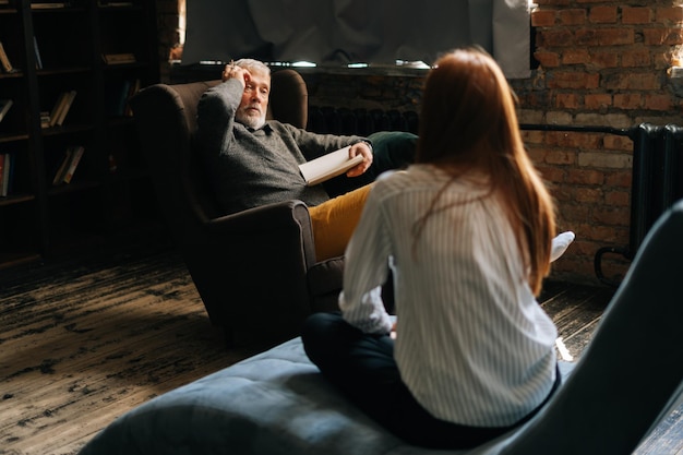 Profesjonalny dojrzały mężczyzna psycholog konsultujący pacjentkę na wizytę psychologiczną w ciemnym biurze domowym Sfrustrowana młoda kobieta siedzi plecami do kamery, słuchając lekarza