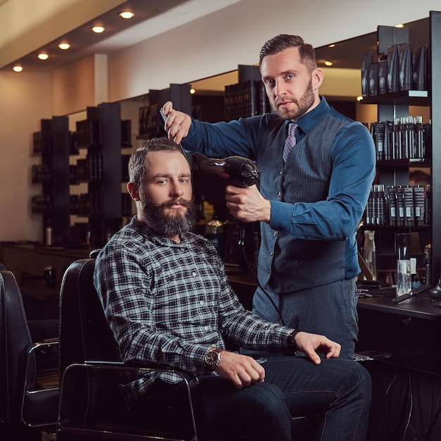 Profesjonalny fryzjer pracujący z klientem w salonie fryzjerskim, korzysta z suszarki do włosów. Strzyżenie męskie w zakładzie fryzjerskim.