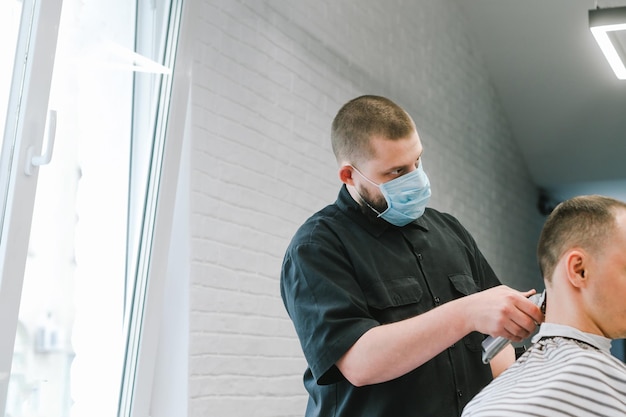 Profesjonalny fryzjer w ochronnej masce z gazy tnie klientce maszynką do strzyżenia w zakładzie fryzjerskim Mężczyzna dostaje fryzurę w zakładzie fryzjerskim podczas kwarantanny fryzjer nosi maskę Koronawirus