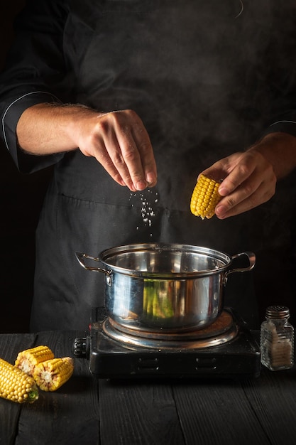 Profesjonalny kucharz dodaje soli do garnka podczas gotowania kukurydzy. Środowisko pracy na vintage kuchennym stole w restauracji. Obraz pionowy.