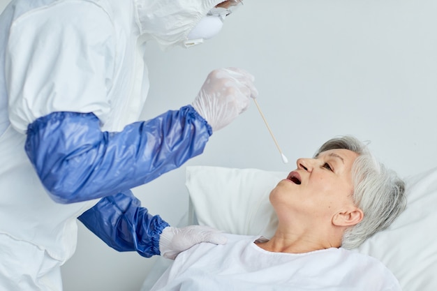 Profesjonalny lekarz w mundurze ochronnym pobierający wymaz starszej kobiety do testu na niebezpieczne wirusy za pomocą sztyftu