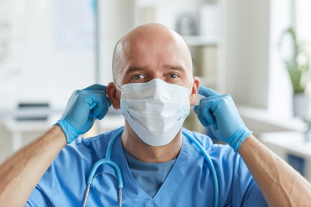Profesjonalny lekarz w niebieskich rękawiczkach mundurowych i lateksowych zakładający maskę ochronną na twarz
