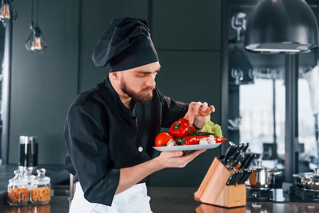 Profesjonalny młody kucharz w mundurze trzyma talerz z warzywami w kuchni