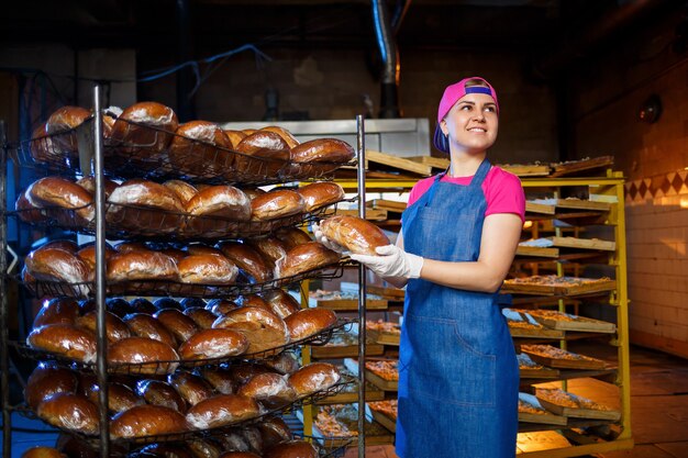 Profesjonalny piekarz - młoda, ładna kobieta w dżinsowym fartuchu trzyma świeży chleb na tle piekarni lub piekarni. Pieczywo. Produkcja chleba