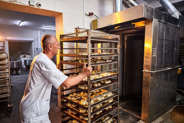 Zdjęcie profesjonalny piekarz w mundurze wkłada wózek z pomostami do wypieku surowego ciasta do wypieku chleba w piecu przemysłowym w piekarni