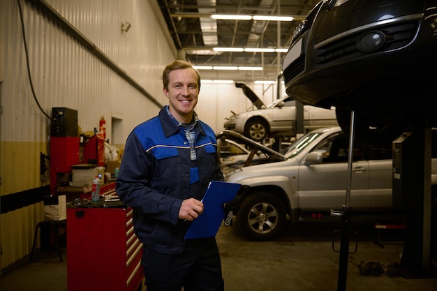 Profesjonalny portret młodego mechanika mechanika samochodowego rasy kaukaskiej w mundurze, uśmiechającego się z uśmiechem zębów, patrzącego na kamerę, stojąc w warsztacie naprawczym ze schowkiem w dłoniach