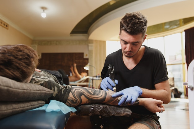 Zdjęcie profesjonalny tatuator wykonujący tatuaż na ramieniu młodego mężczyzny maszynowo czarnym tuszem