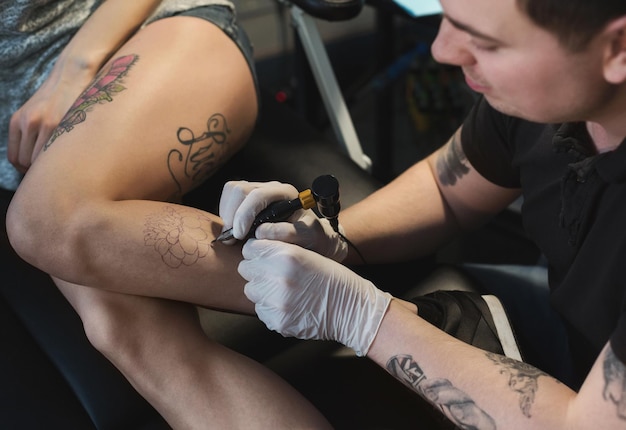 Profesjonalny tatuażysta co piękny tatuaż na nodze młodej kobiety. Mężczyzna tatuażysta rysujący obraz na kobiecym ciele za pomocą specjalnej maszyny, kopia przestrzeń