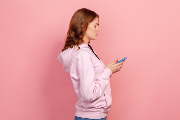 Profil poważne kręcone włosy nastolatka w bluzie z kapturem patrząc na ekran smartfona, czekając na odpowiedź, aplikacja mobilna. Kryty strzał studio na białym tle na różowym tle