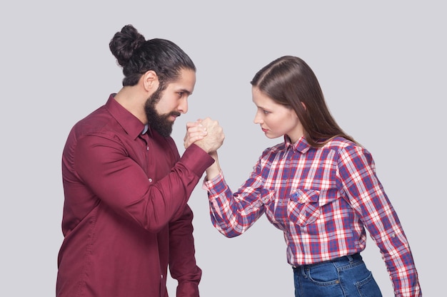 Profil Widok Z Boku Portret Brodaty Mężczyzna I Kobieta W Stylu Casual, Stojąc W Pozie Armwrestlingu I Patrząc Na Siebie Z Poważną Miną. Kryty Strzał Studio, Na Białym Tle Na Szarym Tle.