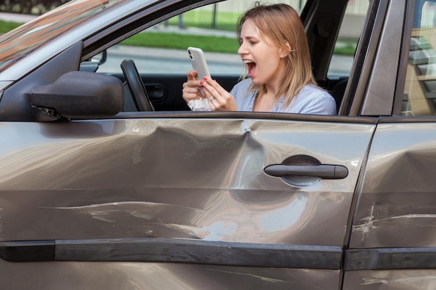 Profil wściekłej, zrozpaczonej kobiety o blond włosach, trzymającej w dłoni inteligentny telefon i głośno krzyczącej podczas dzwonienia do służby ubezpieczeniowej po wypadku drogowym, wgniecenia i zadrapania na drzwiach Zdjęcie na zewnątrz