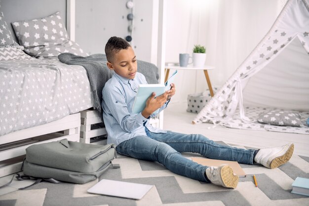 Program szkolny. Atrakcyjny afro-amerykański chłopiec siedzi na podłodze podczas studiowania książki
