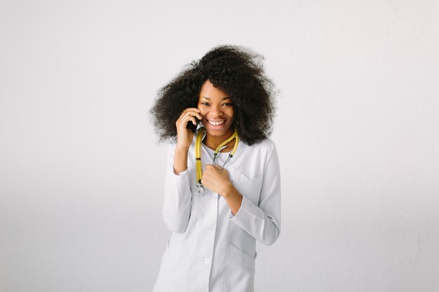 Projekt Reklamy Medycznej. Młoda Piękna Dziewczyna Afroamerykańskiej Dziewczyny W Białym Płaszczu Ze Stetoskopem I Folderu W Szpitalu. Bezosobowe Szerokie Tło. Rozmawiać Przez Telefon