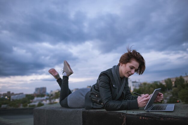 Projektant z tabletem działa na tle zachmurzonego nieba. Kreatywna kobieta leżąca na dachu budynku w mieście podczas pracy. Blogger, praca na odległość, internet bezprzewodowy, koncepcja zawodu artystycznego