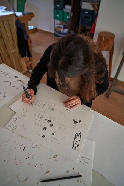 Projektantka bez twarzy wykonująca szkice kreatywnych, ręcznie robionych akcesoriów na kartce papieru w warsztacie jubilerskim pod dużym kątem
