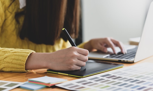 Projektantka szkicuje na cyfrowym tablecie graficznym, Kobieta pisze na cyfrowej tablicy za pomocą pióra i pracuje na laptopie, Biurko projektanta.