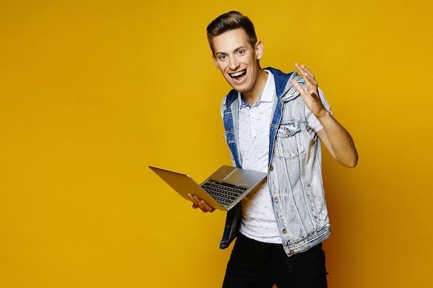 Promieniejący pozytywny facet z laptopem w jego rękach nad żółtym tłem. Młody modniś z notatnikiem odizolowywającym przy żółtym tłem.
