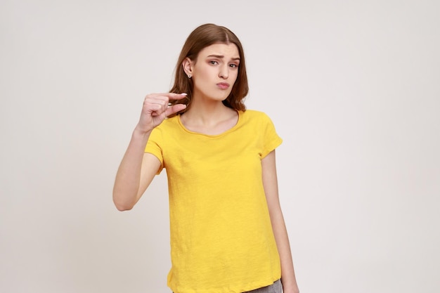 Proszę trochę więcej Portret cute teen dziewczyna w żółtej koszulce pokazujący mały rozmiar lub mały gest palcami patrząc z błagalnym grymasem Kryty studio strzał na białym tle na szarym tle