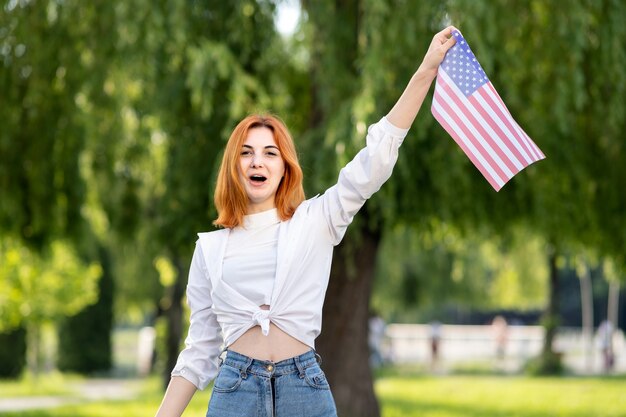 Protestujący zły młoda rudowłosa kobieta pozuje z flagą narodową USA w ręku stojąc na zewnątrz w letnim parku. Aktywistka dziewczyna na demonstracji z narodowym sztandarem.