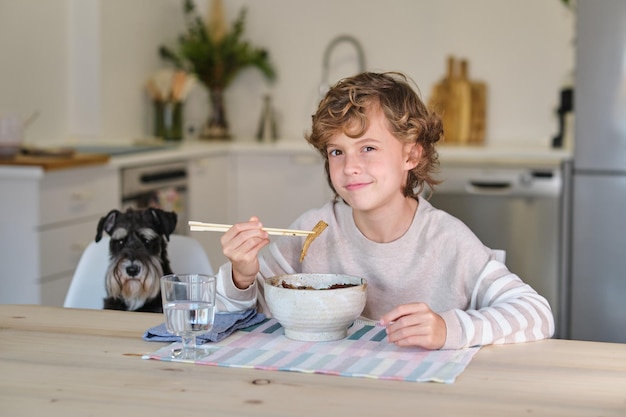 Przebiegłe dziecko siedzi przy drewnianym stole w pobliżu psa i patrzy na kamerę podczas jedzenia smacznego obiadu z pałeczkami w kuchni w domu