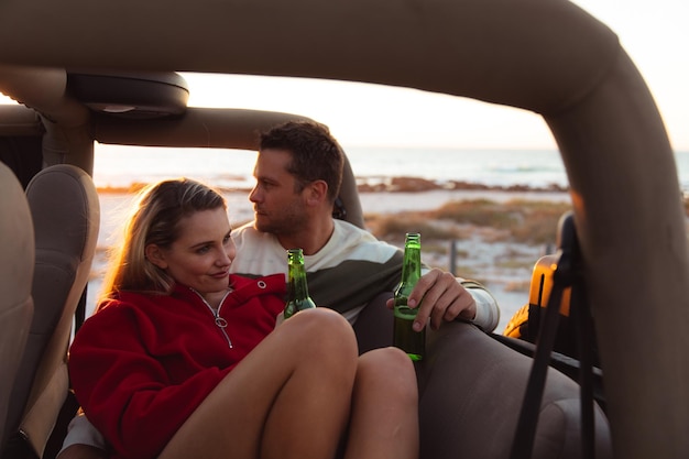 Przedni widok białej pary w samochodzie z otwartym dachem, z zachodem słońca na plaży w tle, uściskając się i trzymając butelki piwa