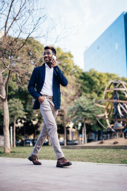 Przedsiębiorca stylowy czarny mężczyzna idący ulicą podczas korzystania z telefonu i robienia interesów