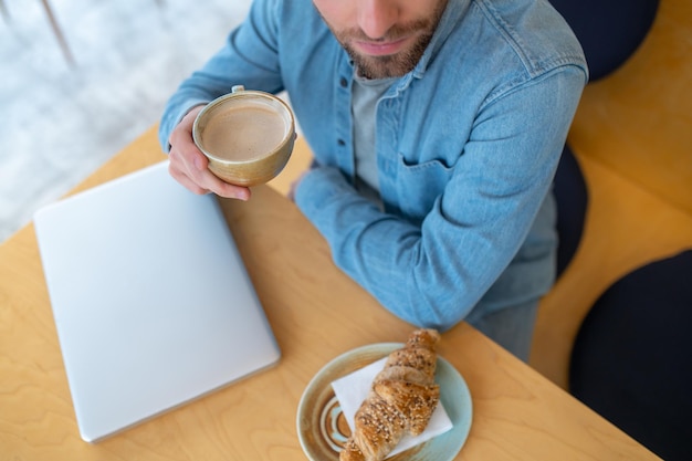Przekąska. Widok z góry człowieka w dżinsowych ubraniach z filiżanką cappuccino siedzącego przy stole z rogalikiem i laptopem w pomieszczeniu