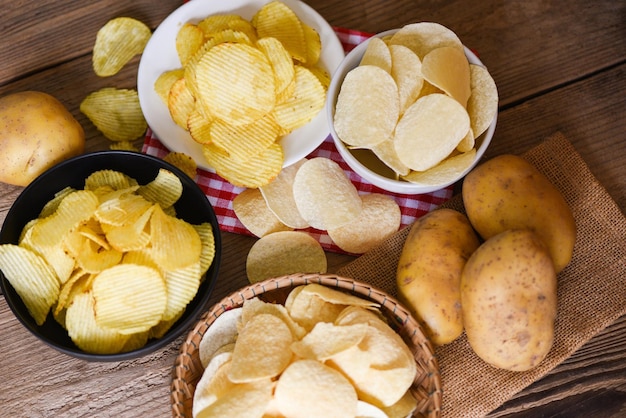 Przekąska z chipsami ziemniaczanymi na misce i talerzu Chrupiące chipsy ziemniaczane na kuchennym stole i świeże surowe ziemniaki na drewnianym tle
