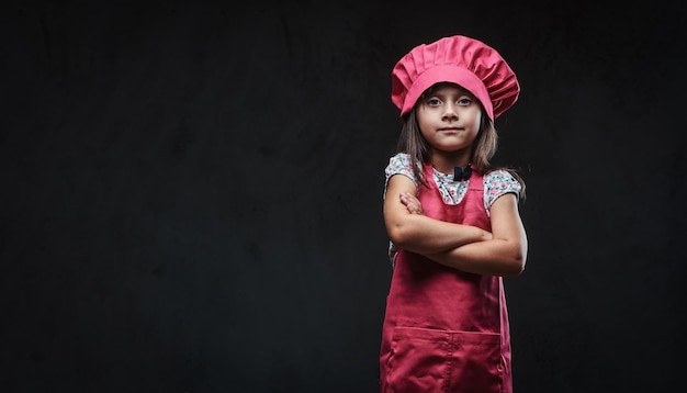 Przekonana, mała dziewczynka ubrana w różowy kucharz jednolity stojący ze skrzyżowanymi rękami. Na białym tle na ciemnym tle z teksturą.