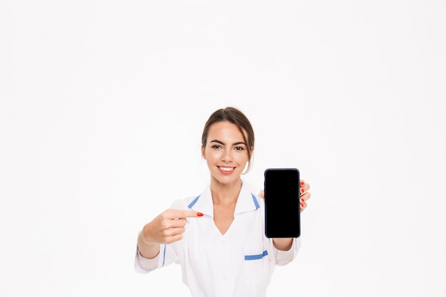 Przekonana, młoda kobieta lekarz ubrany w mundur stojący na białym tle nad białą ścianą, pokazując pusty ekran telefonu komórkowego