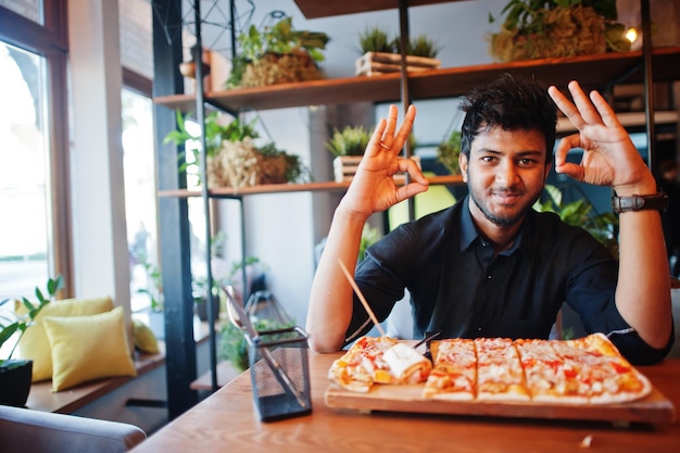 Przekonany, młody indianin w czarnej koszuli, siedzący w pizzerii z pizzą i pokazujący znak ok rękami