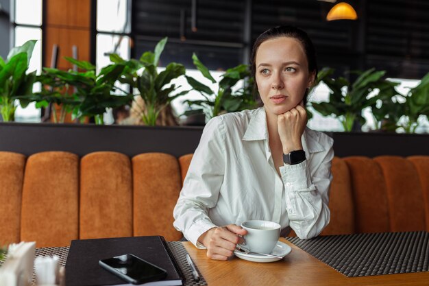 Przemyślana kobieta w białej koszuli pije kawę przy stole w kawiarni