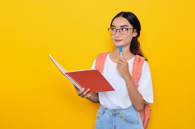 Przemyślana ładna młoda studentka w plecaku i okularach trzyma notatnik i długopis, patrząc na miejsce na kopię na żółtym tle