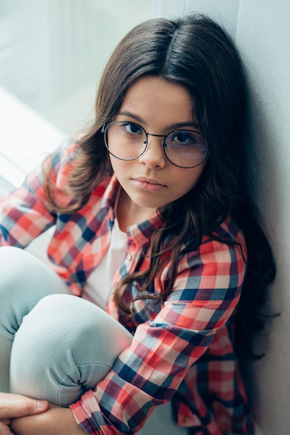 Przemyślana piękna dziewczyna w okularach siedzi pod ścianą i owija ramiona wokół kolan