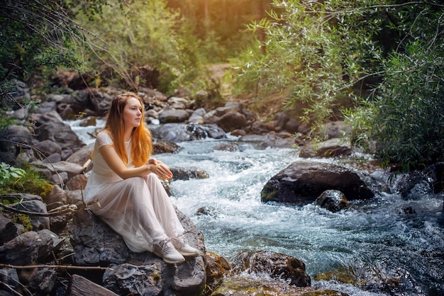 Przemyślana romantyczna młoda dziewczyna w białej sukni siedzi na brzegu górskiej rzeki wśród zielonych drzew. Piękna długowłosa kobieta wypoczywa samotnie z naturą. Koncepcja szczęśliwy styl życia harmonii.