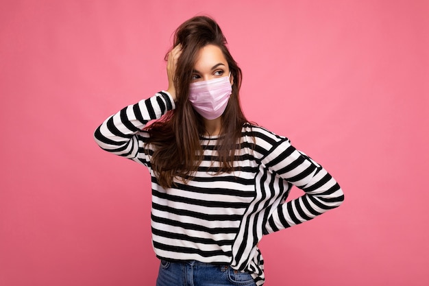 Przemyślane młoda atrakcyjna kobieta nosi maskę medialną na białym tle nad ścianą tła. Ochrona przed poczęciem COVID-19