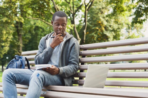 Przemyślany afro-amerykański student studiujący i używający laptopa z notebookiem w parku. Koncepcja technologii, komunikacji, edukacji i pracy zdalnej, kopia przestrzeń