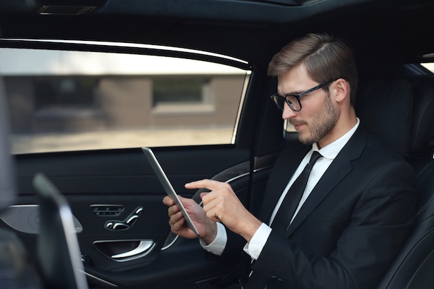 Przemyślany młody biznesmen siedzi w luksusowym samochodzie i za pomocą tabletu.