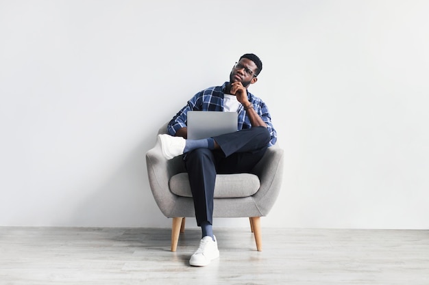 Przemyślany młody czarny człowiek za pomocą laptopa siedząc na krześle na tle białej ściany studia pełnej długości