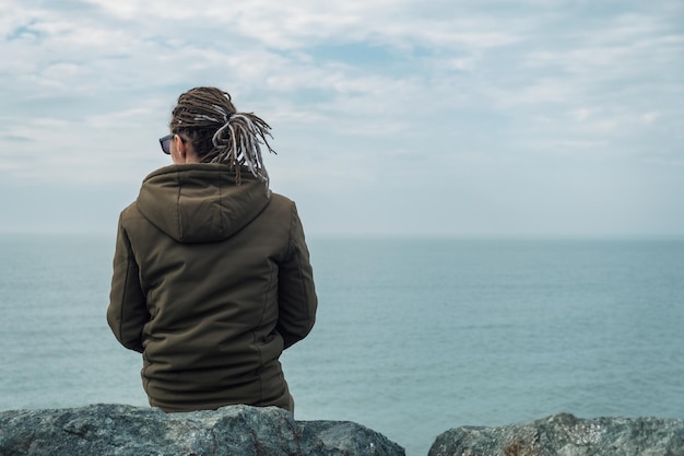 Przemyślany samotny mężczyzna z dredami siedzi nad brzegiem morza w pochmurny dzień Depresja samotność kryzys problemy psychiczne