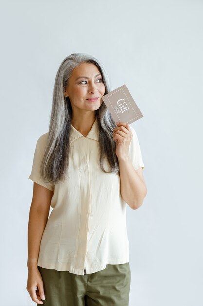 Przemyślany siwy włosach azjatyckich dama klienta posiada kartę upominkową stojącą na jasnoszarym tle w studio. Certyfikat zakupów