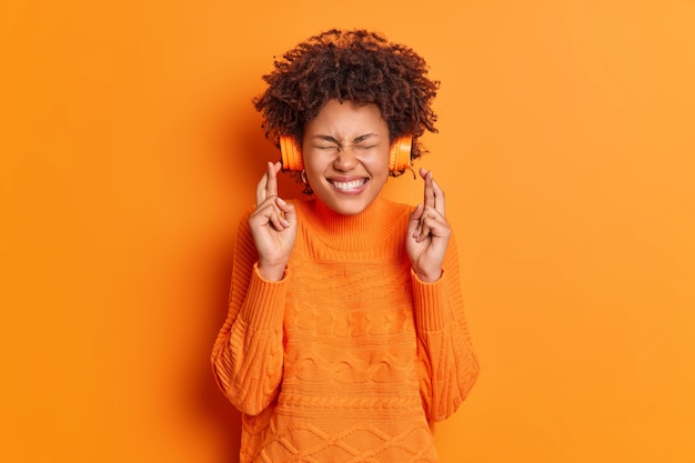 Przesądna młoda, afroamerykańska nastolatka krzyżuje palce, wierzy w szczęście, spełnia życzenia, słuchając muzyki przez słuchawki bezprzewodowe, nosi swobodny sweter odizolowany na pomarańczowej ścianie
