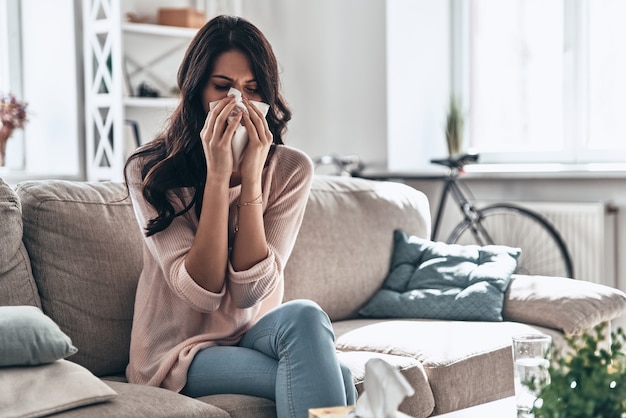 Przeziębienie I Grypa. Chora Młoda Kobieta Wydmuchuje Nos Za Pomocą Bibuły, Siedząc Na Kanapie W Domu