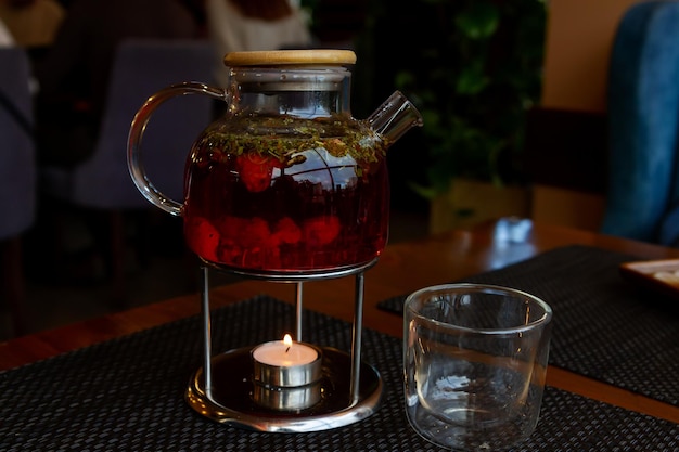 Zdjęcie przezroczysty szklany czajniczek z czerwoną herbatą na podnóżku i świeca stoi na stole obok szkła