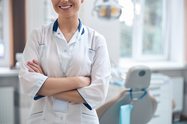Przycięta głowa uśmiechnięta młoda kobieta w mundurze medycznym stojąca w pobliżu fotela dentystycznego i czekająca na pacjentów